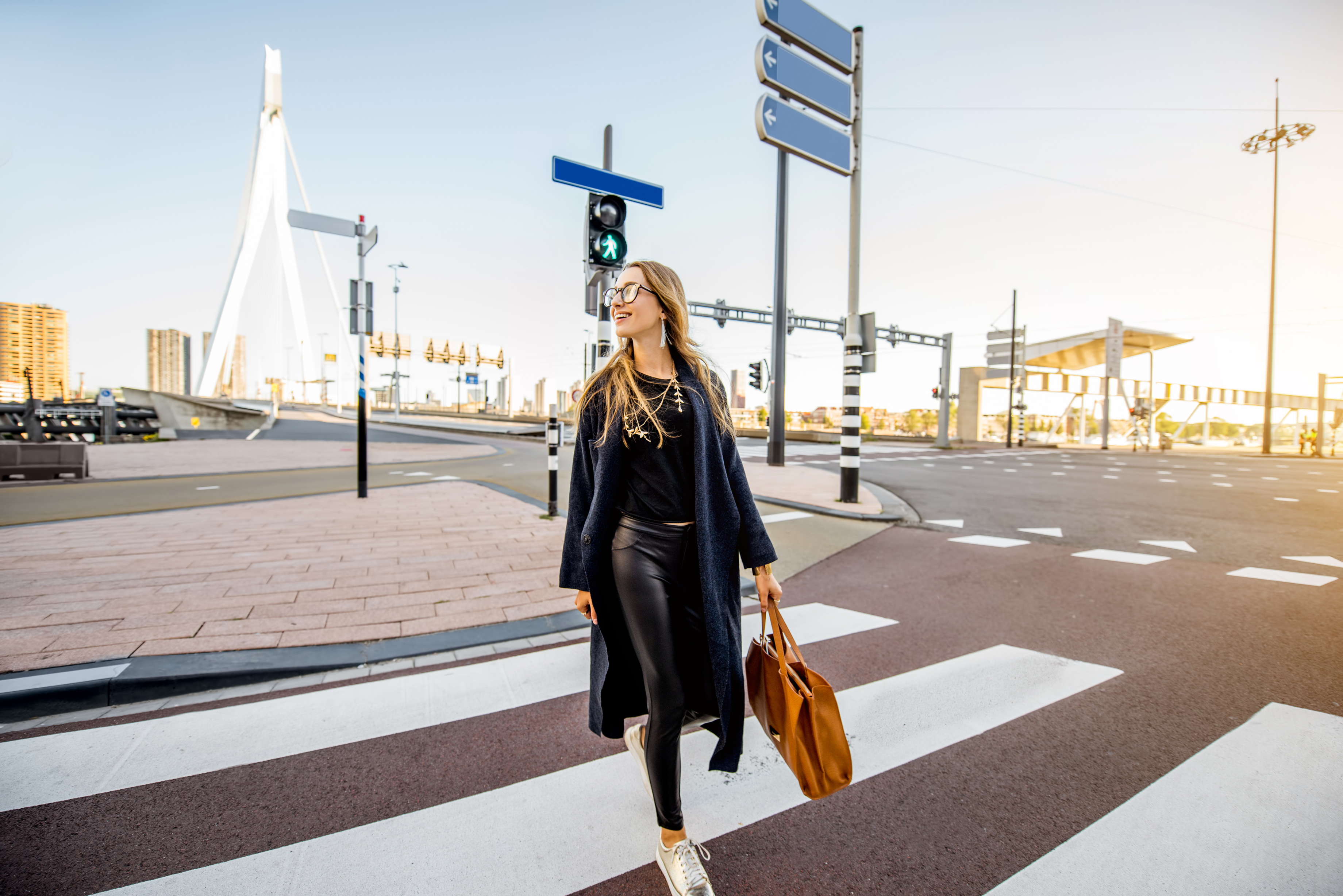 Vrouw met zwarte outfit, bril en bruine tas loopt op een zebrapad naar de camera toe. Het kruispunt ligt voor de Erasmusbrug in Rotterdam, het kruispunt is leeg en de zon schijnt aan de rechterkant in het beeld.