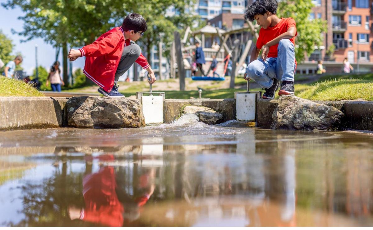 Twee jonge jongens spelen samen met water. Het is relatief warm en de foto is genomen vanaf het perspectief van de grond. We zien de jongens in het midden van het beeld naar elkaar toe zitten en lol hebben met het water.