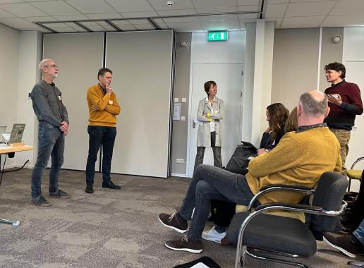 Twee mannen links op de foto zijn in gesprek met iemand uit de zaal, rechts op de foto. Alledrie staan ze, de voorzitter van de meeting staat in het midden de mannen aan te kijken.