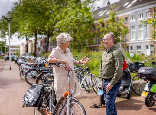 Dame en heer lachen en praten met elkaar. De dame staat met een fiets in de hand. Beiden zijn van middelbare leeftijd. De omgeving is stads met redelijk wat groen.