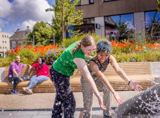 Twee jongvolwassen vrouwen spelen met water bij een fontein aangelegd in Delft. Het is een warme dag. Op de achtergrond zitten nog twee mensen op een lange bank.