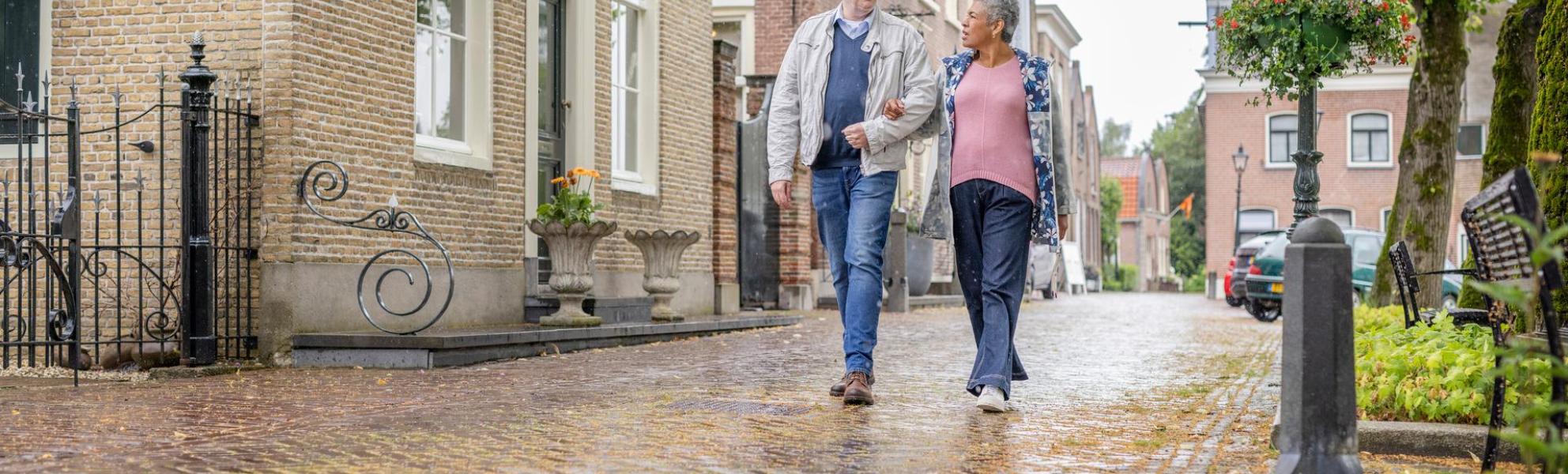 Man en vrouw lopen naast elkaar over straat in Nieuwpoort. Het is een oude straat met mooie woningen en grote bomen. Een voorbeeld van gezonde mobiliteit. De man en vrouw praten met elkaar en lopen op de camera af.