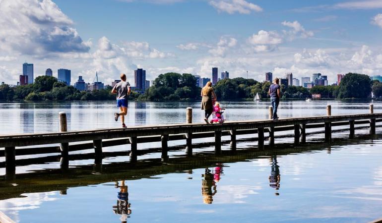 Mensen lopen op een steiger of looppad over het water bij het Kralingse bos in Rotterdam. Je ziet een hardloper, moeder met kindje en een man vanaf de achterkant. Ze lopen dezelfde richting op uit beeld. Op de achtergrond zie je de stad Rotterdam en de Erasmusbrug.