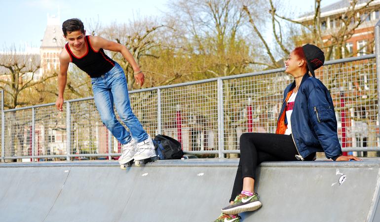 Twee licht getinte jongeren (man, vrouw) staan en zitten op de rand van een skatebaan. De jongen links heeft skeelers aan, de vrouw rechts kijkt lachend naar de man.