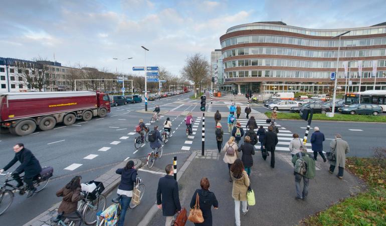 Verkeer en vervoer, kruising ZuidHollandlaan met de Koningskade , in de ochtendspits zijn mensen lopend en fietsend onderweg naar hun werk, autos, motoren, vrachtwagen, bus, vele soorten verkeer komen samen op dit kruispunt, verkeer wacht voor de stoplichten, verkeerslichten reguleren de verkeersstromen, overzichtsfoto over het plein, fietspad, zebrapad, oversteekplaatsen, mensen lopen richting kantoren, provinciehuis, spits in de stad.