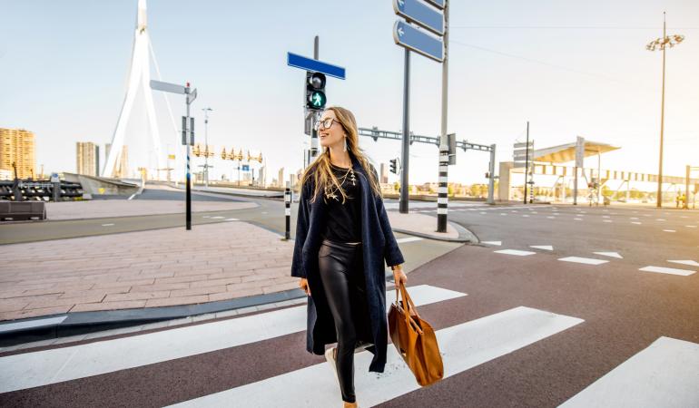 Vrouw met zwarte outfit, bril en bruine tas loopt op een zebrapad naar de camera toe. Het kruispunt ligt voor de Erasmusbrug in Rotterdam, het kruispunt is leeg en de zon schijnt aan de rechterkant in het beeld.