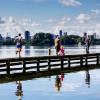 Mensen lopen op een steiger of looppad over het water bij het Kralingse bos in Rotterdam. Je ziet een hardloper, moeder met kindje en een man vanaf de achterkant. Ze lopen dezelfde richting op uit beeld. Op de achtergrond zie je de stad Rotterdam en de Erasmusbrug.