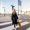 Vrouw met zwarte outfit, bril en bruine tas loopt op een zebrapad naar de camera toe. Het kruispunt ligt voor de Erasmusbrug in Rotterdam, het kruispunt is leeg en de zon schijnt aan de rechterkant in het beeld.